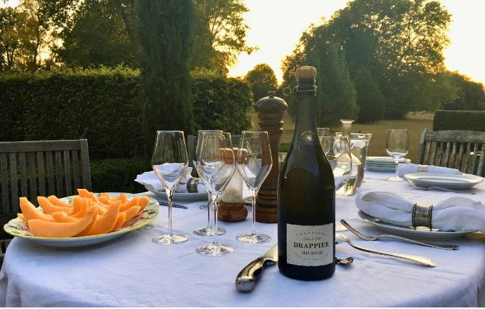 table dressée dehors dans le parc avec verres de champagne, melon en tranche, soleil couchant sur le parc